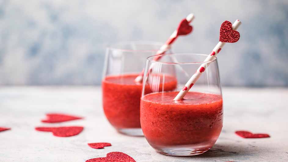 Twee glazen met rode smoothies en gestreepte rietjes met hartjes. Losse rode hartjes liggen verspreid op het oppervlak.