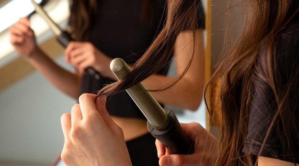 Person styling long brown hair with a curling iron in a well-lit room, wearing black clothing.