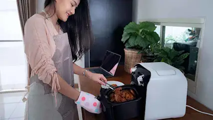 Vrouw met ovenwant en tang haalt eten uit een witte airfryer. Op de achtergrond een laptop en planten.
