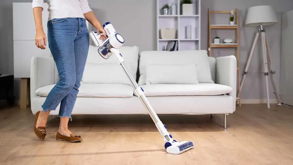 Vrouw stofzuigt houten vloer met een witte steelstofzuiger in een woonkamer met witte bank en kast.