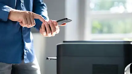 Een persoon met een blauw overhemd gebruikt een smartphone naast een zwarte printer in een kamer met een raam.