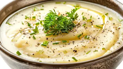 Een kom met aardappelpuree, gegarneerd met peterselie, bieslook en zwarte peper. Er is een scheutje olie op de puree.