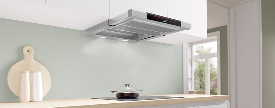 Een keuken met een zilverkleurige afzuigkap boven een kookplaat met een pan erop. Een houten snijplank en witte vazen staan op het aanrecht.