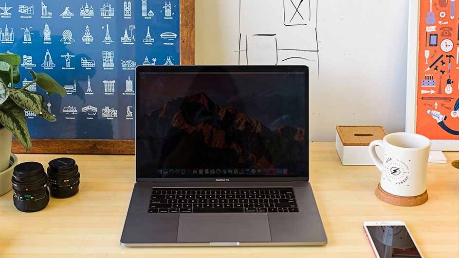 A desk with a laptop, lenses, mug, and phone. There's framed art and a plant on the desk, with a world monuments poster behind.