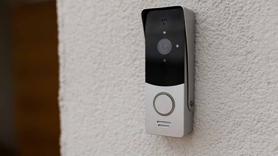 Een rechthoekige, zilverkleurige slimme deurbel met een zwarte bovenkant is aan een witte muur bevestigd. De deurbel heeft een camera en een belknop.