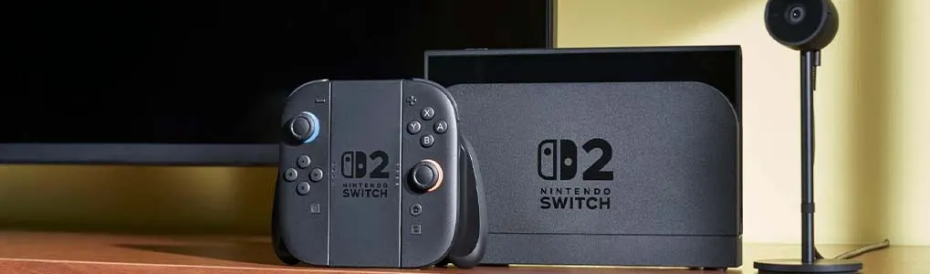 A gray Nintendo Switch OLED console and controller sit on a wood surface. A black television and microphone are in the background.