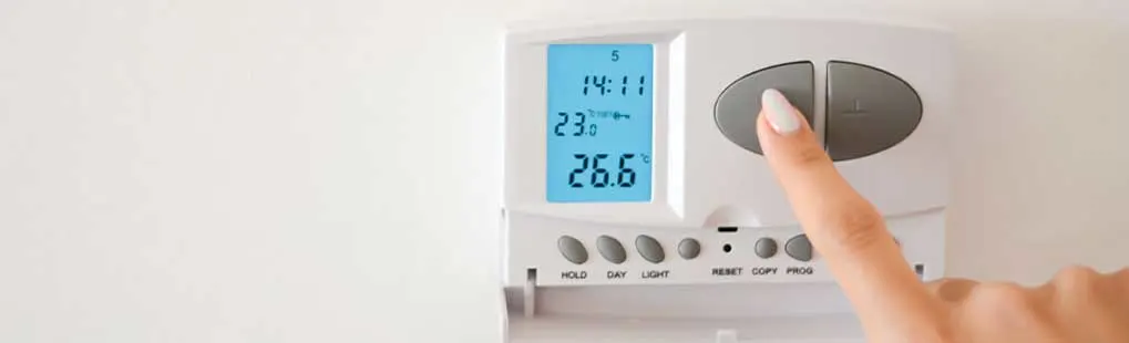 A white thermostat displays 14:11 and 26.6 degrees. A finger presses a grey button on the thermostat.