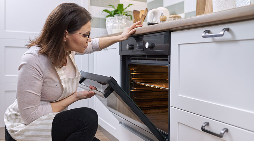 Een vrouw met een schort opent een zwarte oven in een witte keuken.