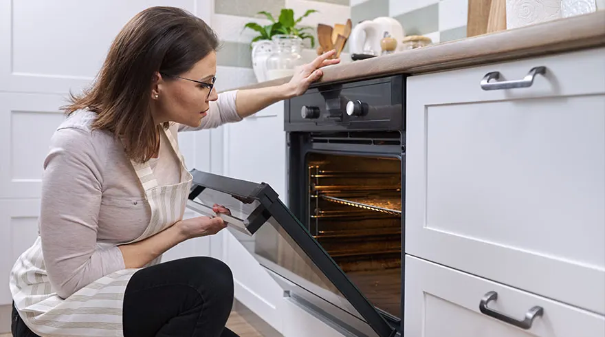 Een vrouw met een schort opent een zwarte oven in een witte keuken.
