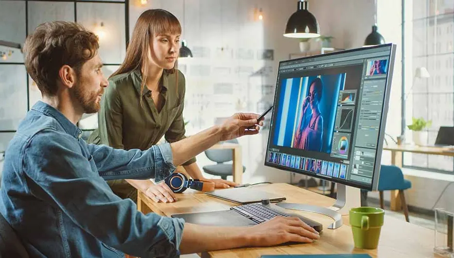 Two people are at a desk. One points to a computer screen showing photo editing software. Headphones and a green mug are on the desk.