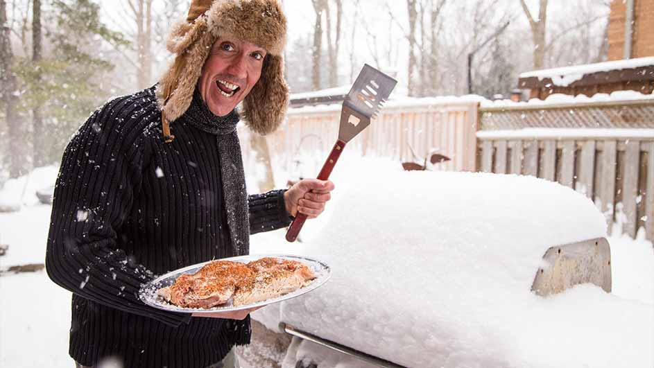 Man met bontmuts houdt bord met vlees en spatel vast bij een besneeuwde barbecue in een winterse omgeving.