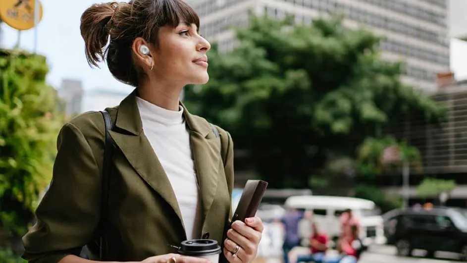 Een vrouw met oortjes, een wit shirt en een groen jasje houdt een telefoon en een koffiebeker vast in een stedelijke omgeving.