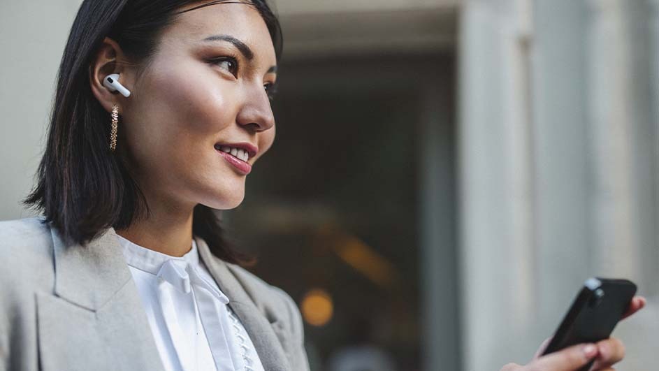 A person with short black hair wears a light-gray blazer and white shirt, smiling and holding a black smartphone.  She has earbuds. Background shows gray walls.