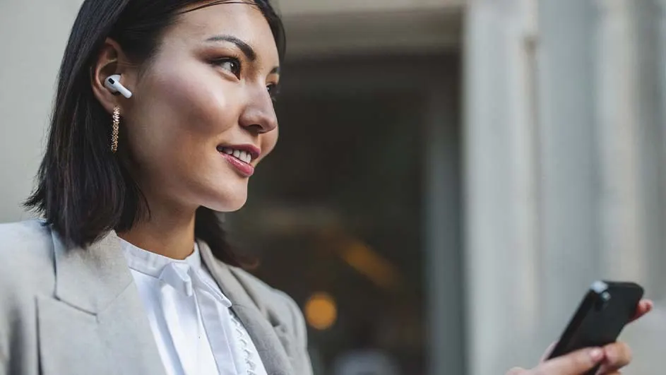 A person with short black hair wears a light-gray blazer and white shirt, smiling and holding a black smartphone. She has earbuds. Background shows gray walls.