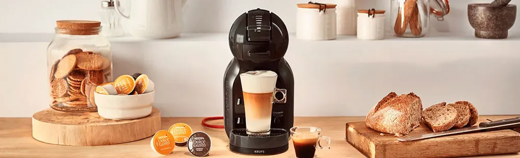 In een keuken staat een zwarte KRUPS NESCAFÉ Dolce Gusto koffiemachine met koffie en capsules, omringd door brood en koekjes.