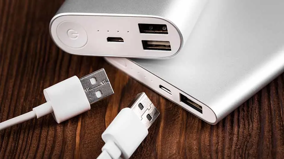 Twee zilverkleurige powerbanks en witte USB-kabels liggen op een houten tafel. De powerbanks hebben verschillende poorten.