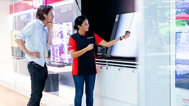 Verkoopster in rood-zwart shirt demonstreert een tv aan een klant in een winkel. Er staan meerdere tv's op een rij.