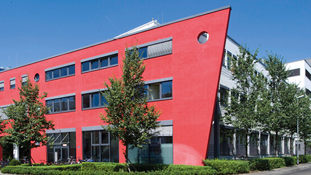 Rood en grijs kantoorgebouw met meerdere ramen en bomen tegen een blauwe lucht.