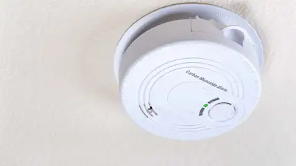 Een witte koolmonoxidemelder is aan een wit plafond bevestigd, met een groen lampje dat brandt.