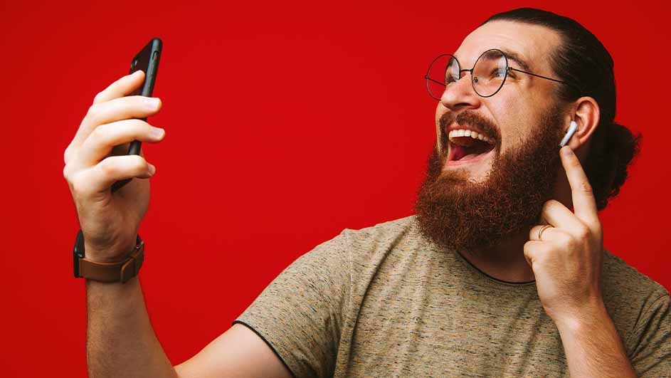 A person with glasses and a beard is smiling and holding a phone. They have an earbud in one ear and are wearing a watch. The background is red.