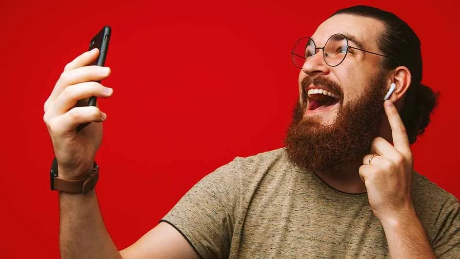 A person with glasses and a beard is smiling and holding a phone. They have an earbud in one ear and are wearing a watch. The background is red.