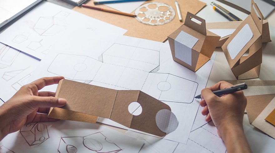 Ontwerp van kartonnen verpakkingen op een witte tafel met handen, potloden en prototypes.