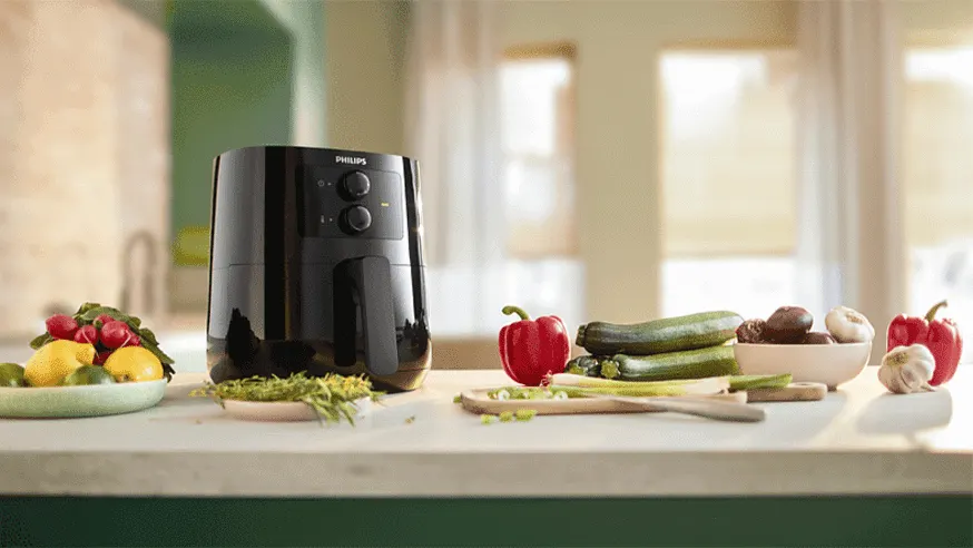 A black Philips air fryer sits on a counter with vegetables, including zucchini, peppers, garlic, lemons, and radishes.