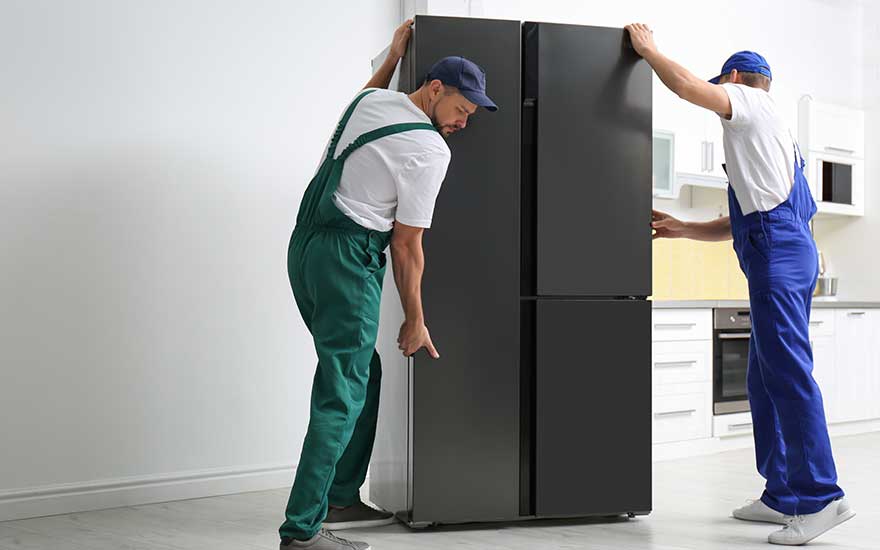 Two technicians in green and blue workwear position a tall black fridge upright in a bright, modern kitchen setting.