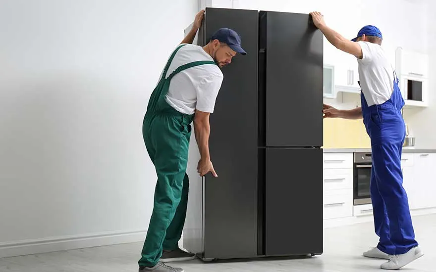 Two technicians in green and blue workwear position a tall black fridge upright in a bright, modern kitchen setting.