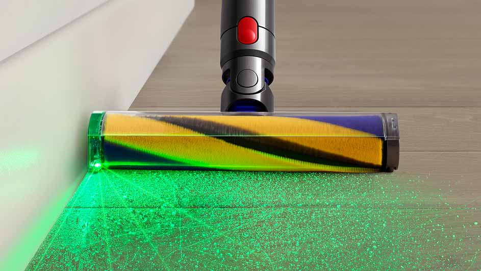 Een stofzuiger met een borstel met gele en blauwe strepen reinigt een houten vloer met groen licht dat stof onthult.