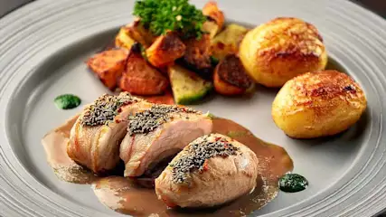 Op een grijs bord liggen stukken kip met kruiden, aardappelen en groenten. Er is een bruine saus bij het vlees.