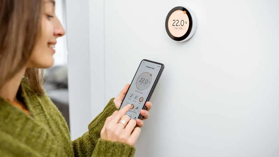 Vrouw bedient een thermostaat met een smartphone. Aan de muur hangt een ronde thermostaat. De temperatuur is 22 graden.