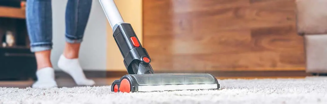 A person vacuums a soft carpet with a modern vacuum cleaner; only the legs are visible and the floor looks fresh and neat.