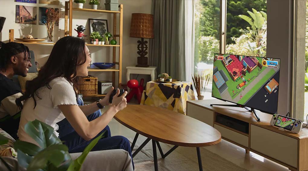 Twee mensen spelen een videospel op een groot scherm in een woonkamer met een houten tafel en een plant op de voorgrond.
