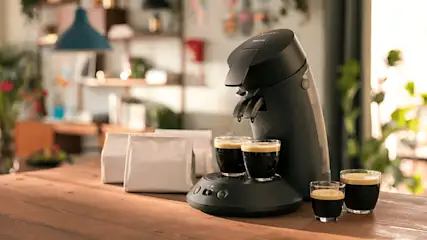 Een zwarte koffiemachine met twee kopjes espresso, witte zakken koffie en extra espresso op een houten tafel.