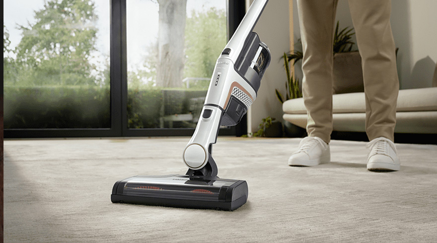 Een staande stofzuiger staat op een beige tapijt, een persoon staat erachter met witte schoenen.