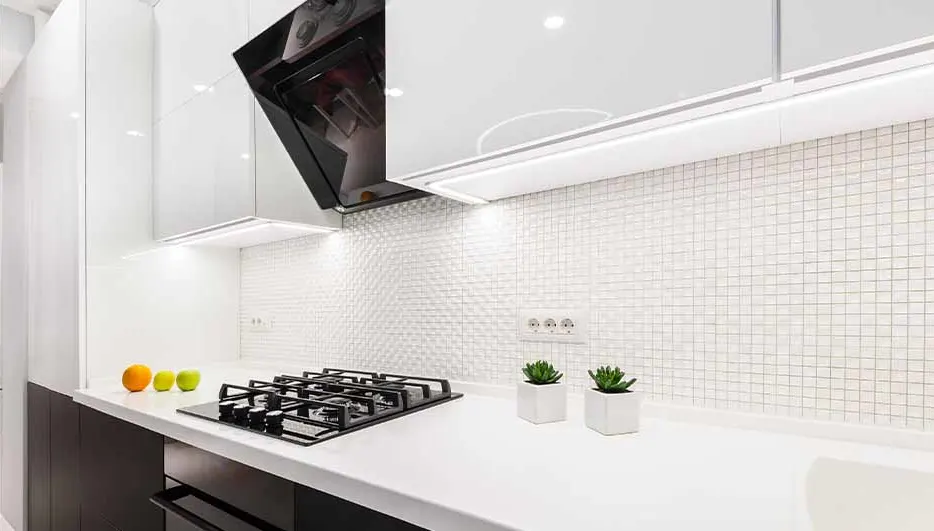 Een keuken met witte kasten, een zwart fornuis, en een witte achterwand met kleine tegels. Er staan twee planten en fruit op het aanrecht.