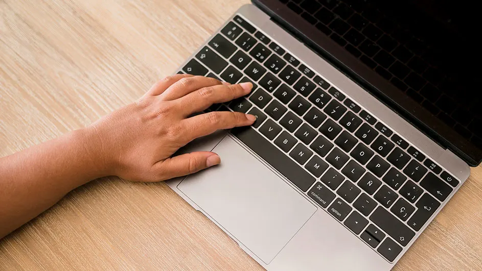 Hand typt op een zwart toetsenbord van een open zilverkleurige laptop op een houten tafel.