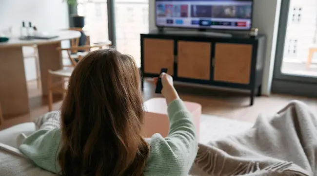 A woman sits on a couch with a blanket, holding a remote control, facing a TV on a cabinet, with a window in the background.