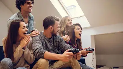 Een groep van zes mensen zit op een bank, spelend met controllers, terwijl ze naar een scherm kijken.