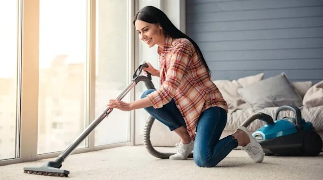 A person in a plaid shirt and jeans vacuums near a window. A blue vacuum cleaner sits on the carpet next to a couch with pillows.