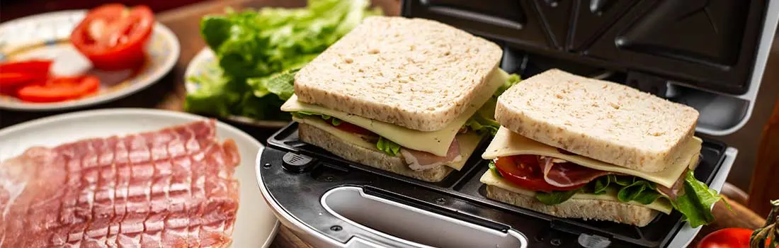 Open sandwich maker met two untoasted sandwiches, surrounded by fresh lettuce, tomato, ham and cheese on a wooden worktop, ready to grill.