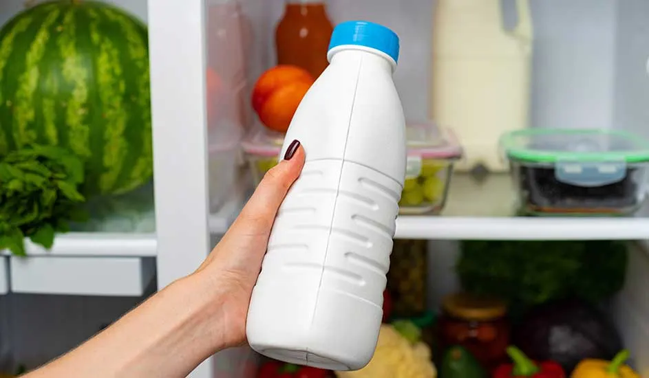 Een hand houdt een witte fles met blauwe dop voor een open koelkast gevuld met een watermeloen, groenten, fruit en bakjes met eten.