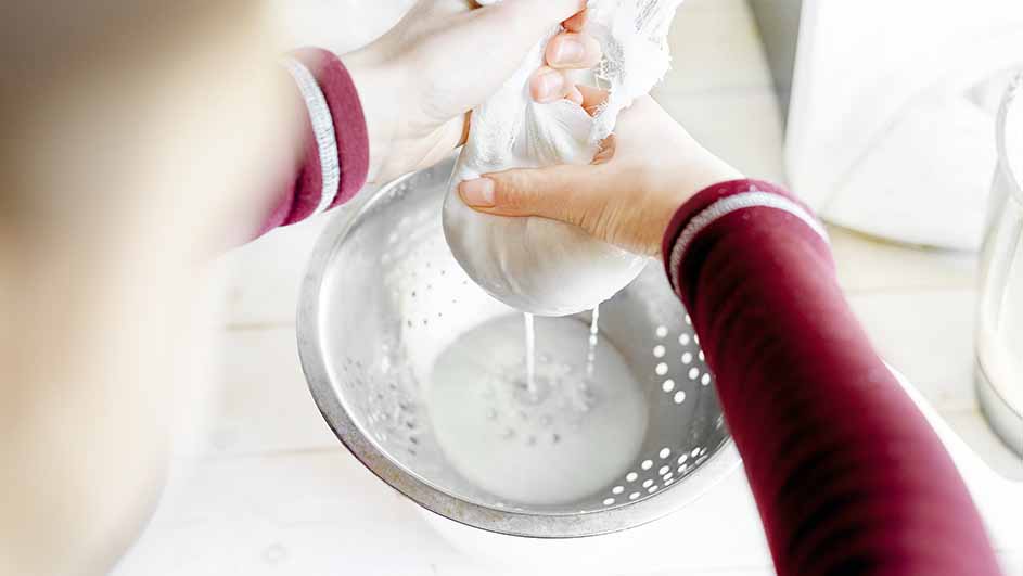 Twee handen persen een witte doek uit boven een metalen vergiet gevuld met vloeistof.