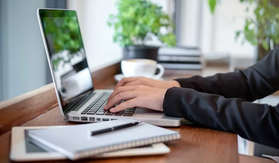 Op de afbeelding typt iemand op een zilveren Apple MacBook laptop aan een houten bureau, met een notitieboek, pen en planten op de achtergrond.