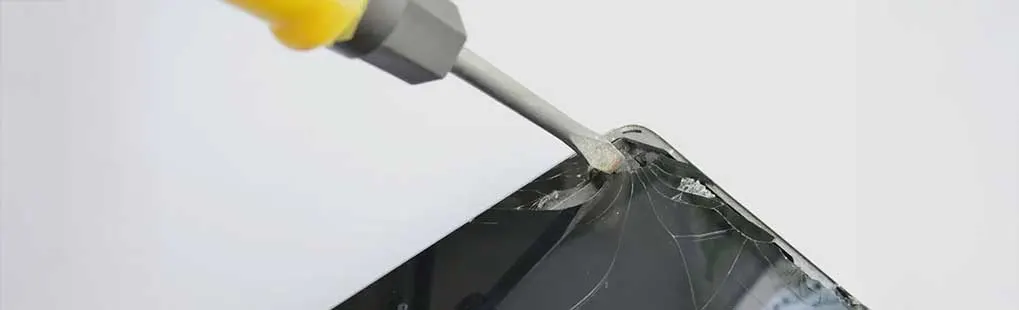 A yellow and gray screwdriver pries open a cracked black electronic device against a plain white backdrop.