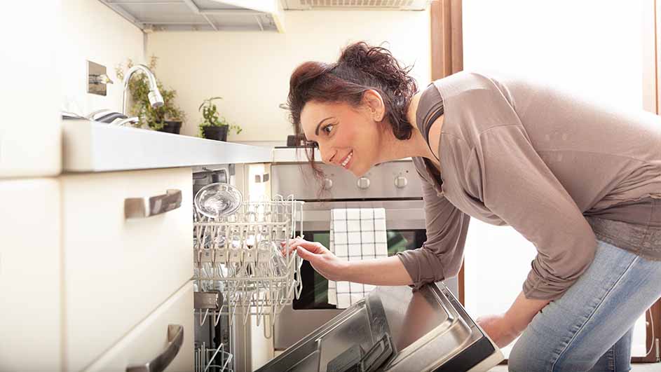 Een vrouw in een keuken laadt een vaatwasser in, omringd door witte kasten en een oven.