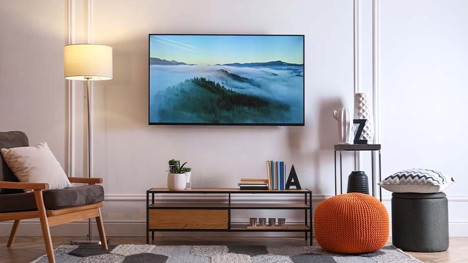 Living room view with a TV showing landscape, chair, lamp, and shelves with books and decorations on a patterned rug.