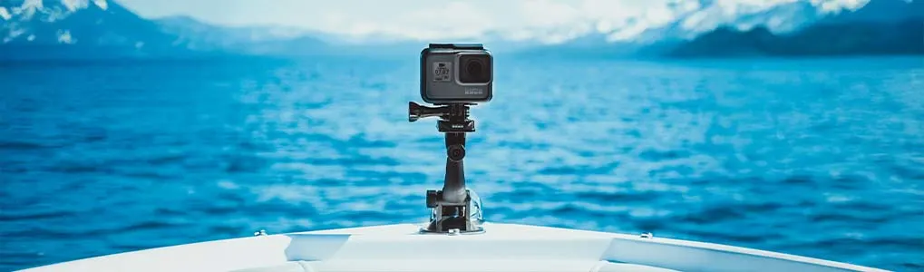 Een scherp gefotografeerde GoPro-camera, bevestigd op de boeg van een boot, met op de achtergrond een helderblauwe zee en besneeuwde bergen in de verte.
