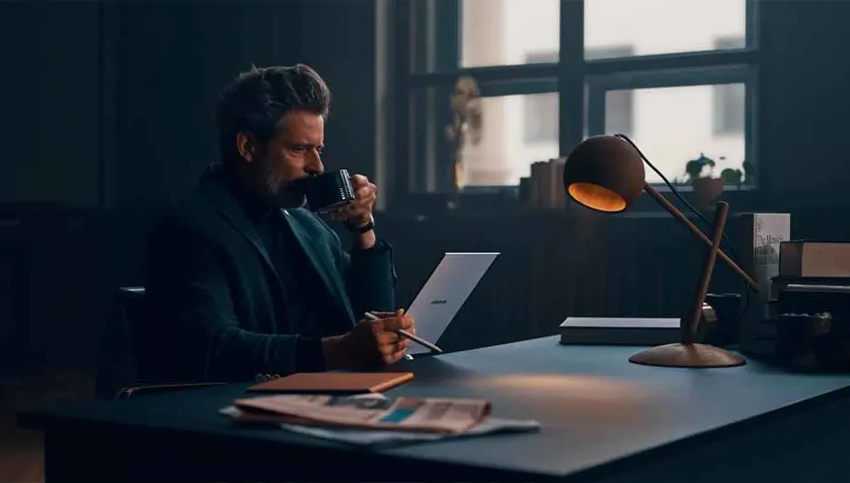 Een man zit aan een bureau met een laptop en een lamp, en drinkt uit een kopje. Kranten liggen op het bureau.